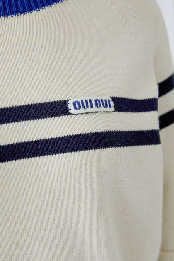 Close-up of a cream-colored knit sweater with navy blue horizontal stripes and a small white brand patch reading 'OUI OUI' on the chest.