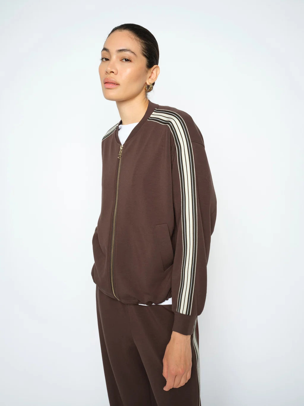 A model wearing a dark brown zip-up track jacket with white and black stripes down the sleeves, paired with matching brown pants, against a plain light gray background.