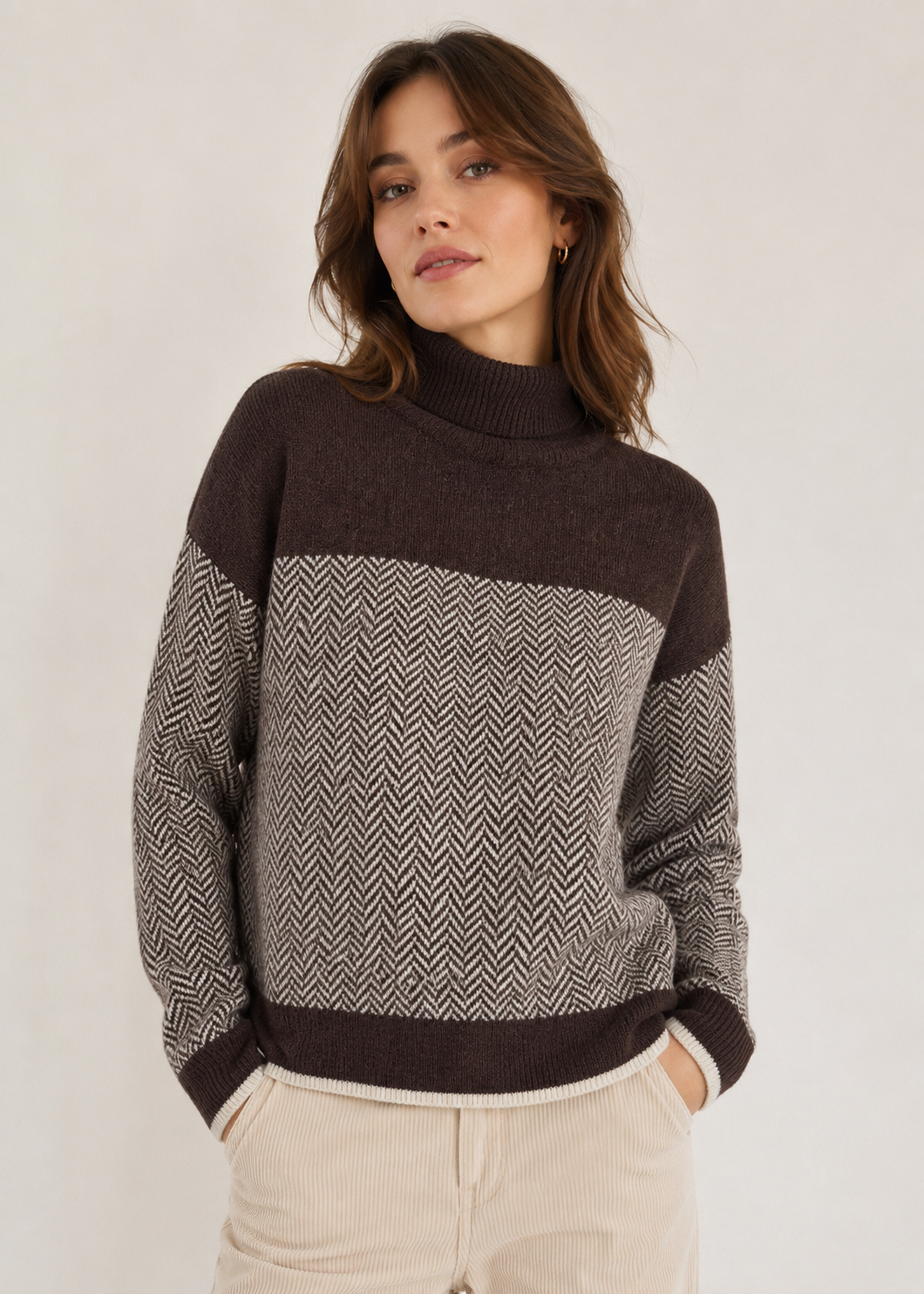 A woman wearing a brown and gray color-blocked knit sweater with a herringbone pattern, paired with light beige corduroy pants, against a plain off-white background.