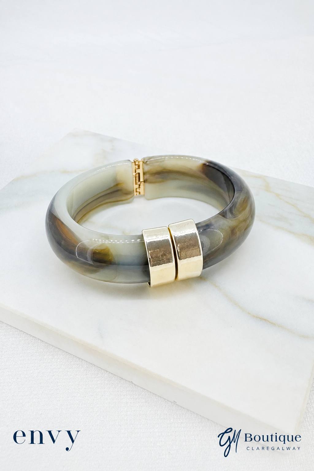 GM Boutique A chunky bangle bracelet with a marbled translucent resin body in shades of gray, brown, and cream, accented by two polished gold magnetic clasp ends, displayed on a white marble slab.