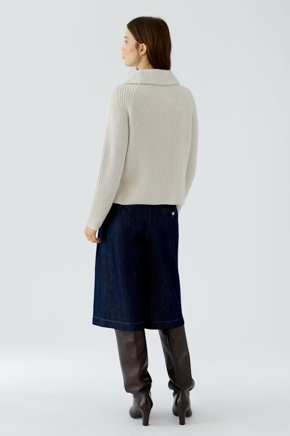 A woman is shown from the back wearing a cream ribbed turtleneck sweater, a dark blue denim midi skirt, and dark brown leather knee-high boots against a light gray studio background.