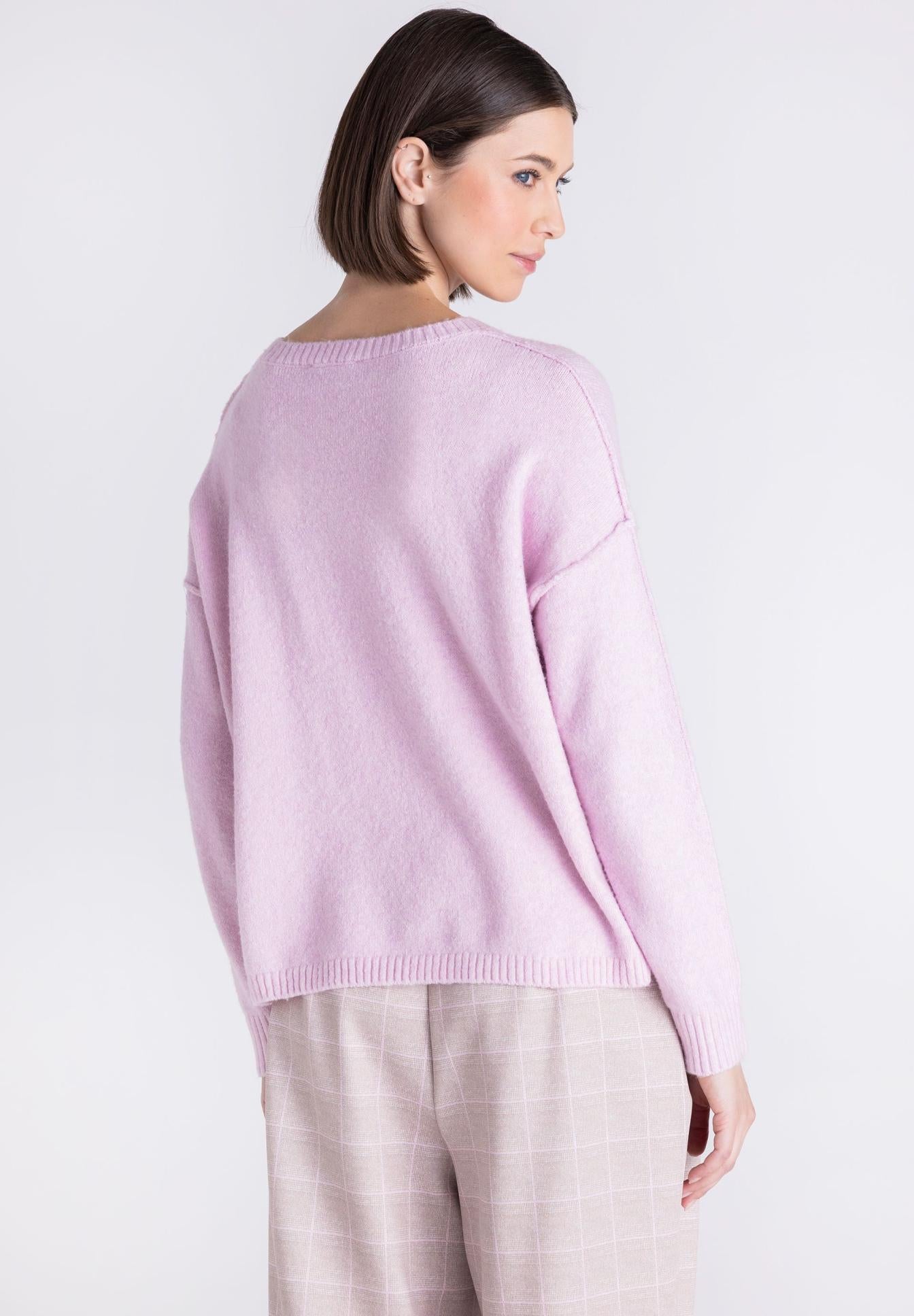 A woman shown from the back wearing a light pink knit sweater and light beige plaid trousers against a plain light gray studio background.