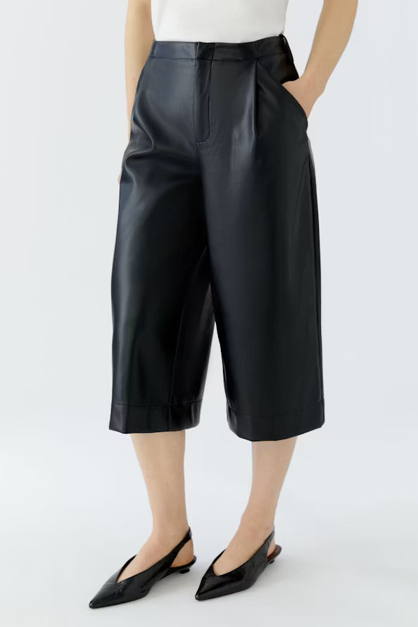A model wearing black faux-leather cropped culotte pants with a high waist, pleated front, and side pockets, paired with a white top and black pointed slingback flats, against a light gray studio background.