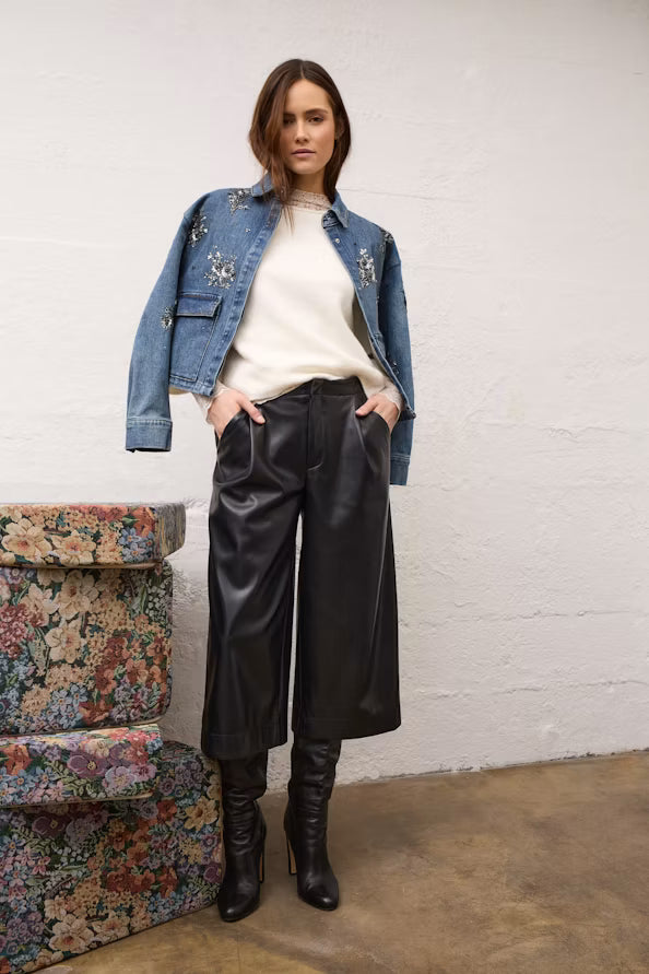 A woman stands indoors wearing a light blue denim jacket with silver embroidery, a cream ribbed top, black wide-leg cropped pants, and black knee-high boots. She is posed in front of a white wall with a floral-patterned bench on the left.