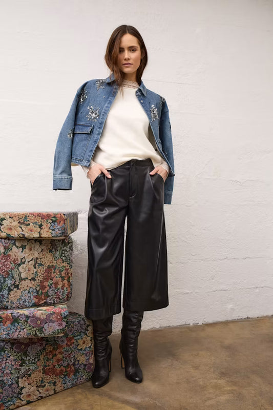 A woman stands indoors wearing a light blue denim jacket with silver embroidery, a cream ribbed top, black wide-leg cropped pants, and black knee-high boots. She is posed in front of a white wall with a floral-patterned bench on the left.