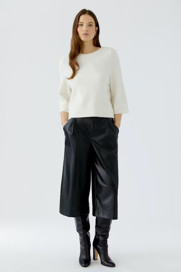 A woman modeling a cream knit sweater and black wide-leg cropped pants with black knee-high boots, photographed against a light gray studio background.