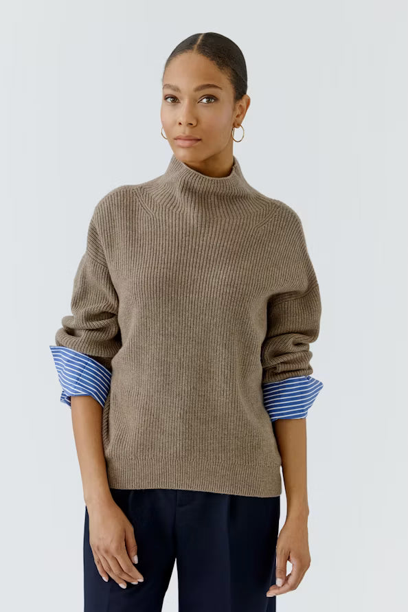 A woman modeling a ribbed knit turtleneck sweater in taupe, with rolled-up sleeves showing blue and white striped cuffs, paired with dark navy trousers against a light gray studio background.