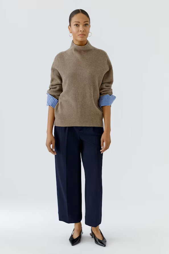 A model stands against a light gray studio background wearing a taupe-brown ribbed knit mock-neck sweater with blue and white striped cuffs, navy wide-leg cropped trousers, and black pointed flats.
