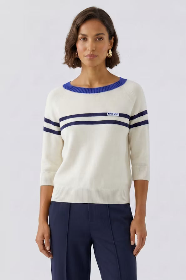 A woman modeling a cream knit sweater with navy blue stripes and a blue crew neck, paired with navy trousers, against a light gray studio background.