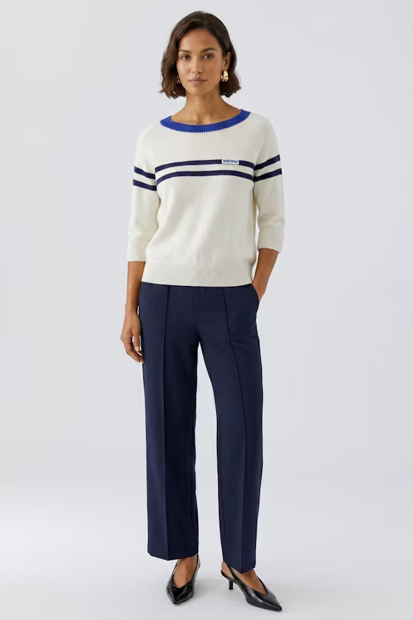 A woman modeling a cream knit sweater with blue and navy stripes, paired with navy trousers and black pointed-toe heels, against a light gray studio background.