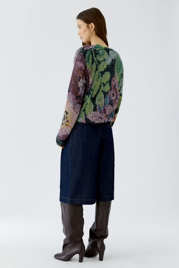 A model is shown from the back wearing a colorful, patterned knit cardigan with green, purple, and pink tones, paired with dark blue denim culottes and dark brown knee-high boots, against a plain light gray studio background.