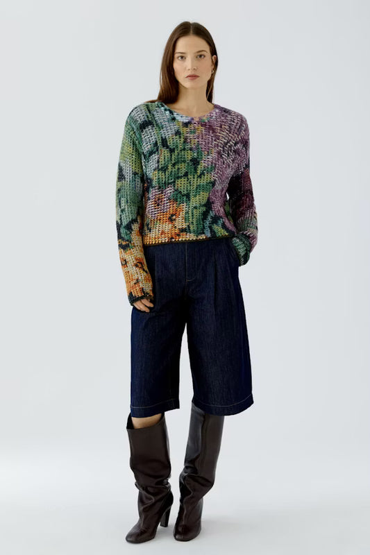 A model stands against a light gray studio background wearing a multicolor patterned knit sweater, dark blue wide-leg cropped jeans, and dark brown knee-high boots.