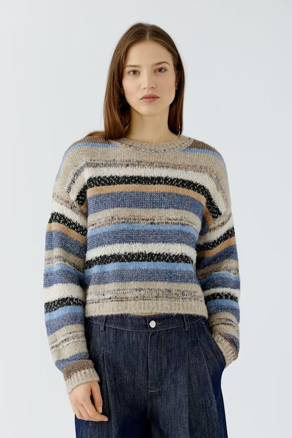 A woman wearing a fuzzy striped knit sweater with horizontal bands of beige, blue, brown, and black, paired with dark blue jeans, against a light gray studio background.