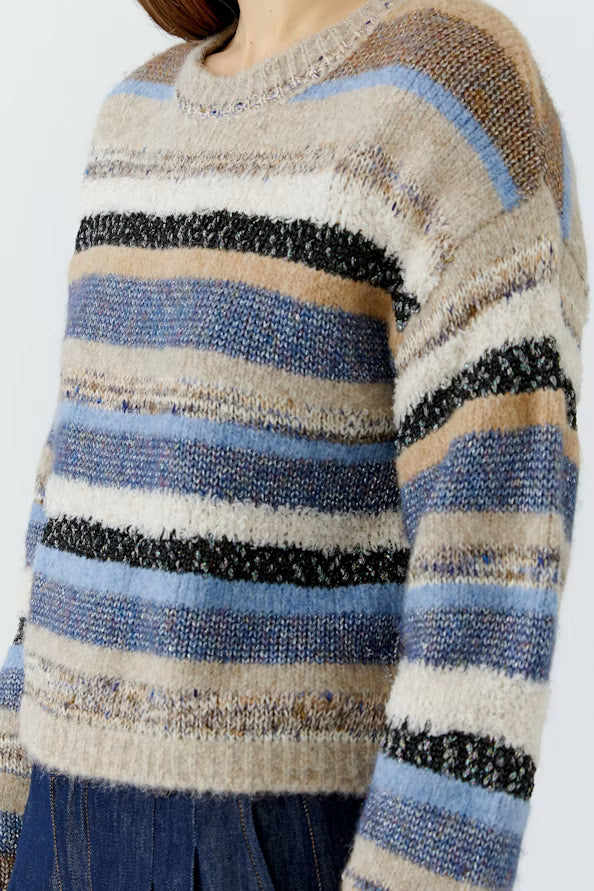 A close-up of a fuzzy knit sweater with horizontal stripes in beige, light blue, medium blue, black, and tan, worn by a model with blue jeans visible at the bottom.