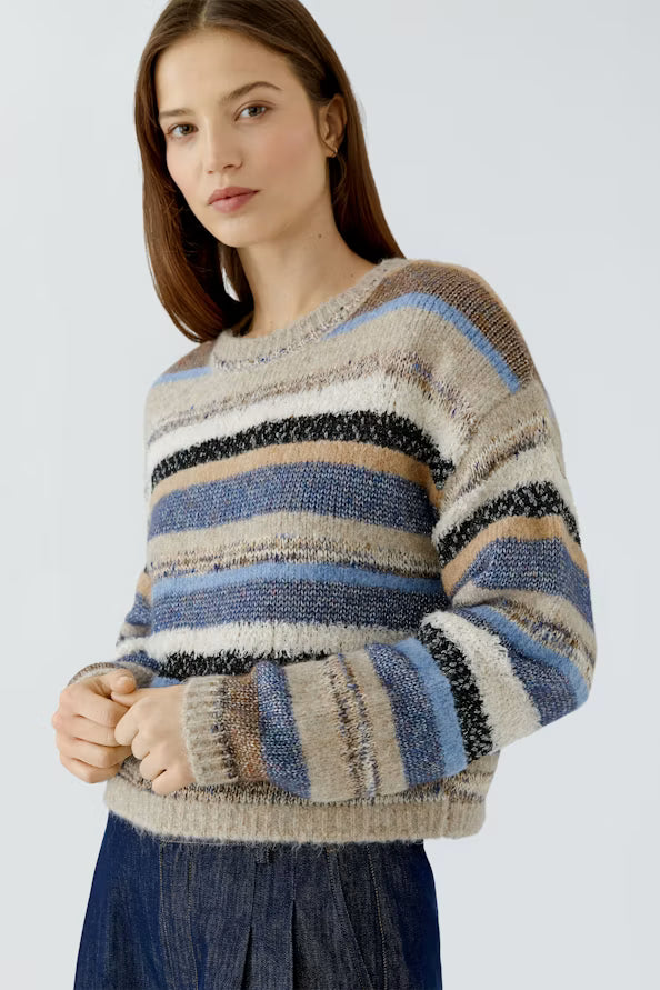 A woman wearing a fuzzy, striped knit sweater with horizontal bands of beige, brown, blue, black, and white, paired with a dark blue skirt, against a light gray studio background.