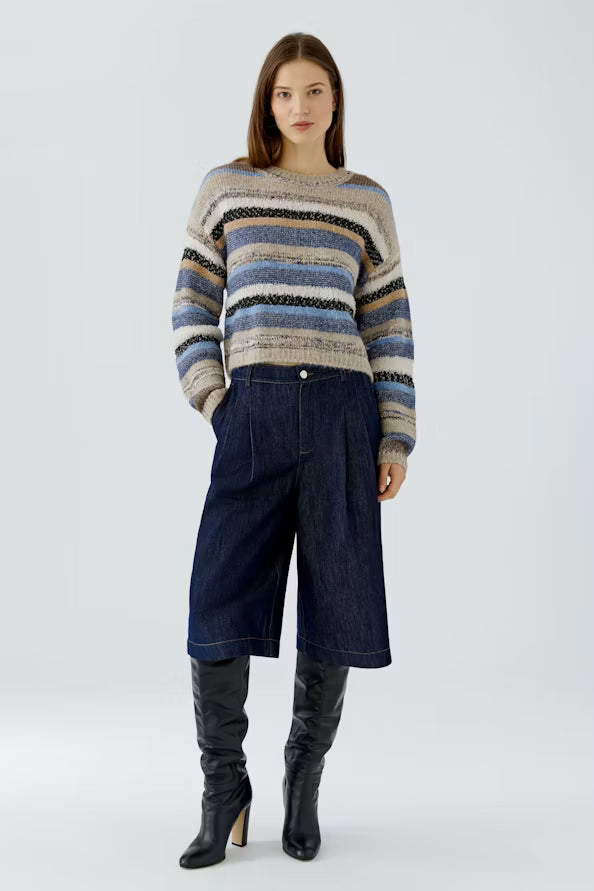 A model stands against a light gray studio background wearing a knit sweater with horizontal stripes in beige, blue, and black, paired with dark blue wide-leg denim shorts and black knee-high boots.