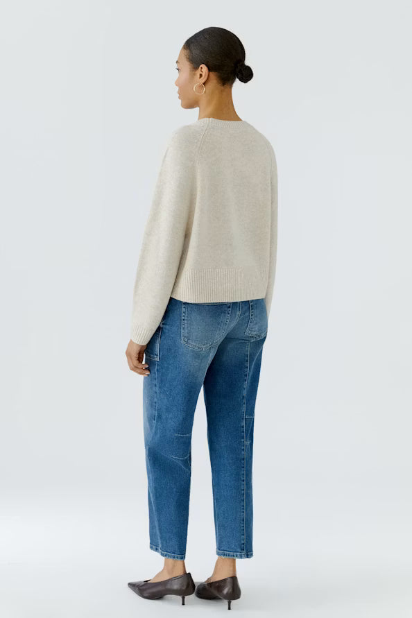 A woman shown from the back wearing a light beige knit sweater, blue cropped jeans, and dark brown pointed-toe heels against a plain light gray studio background.