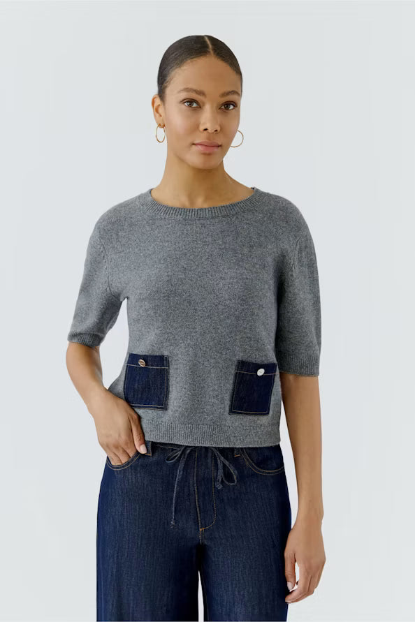 A woman modeling a gray short-sleeve knit sweater with two dark blue denim-style front patch pockets, paired with dark blue jeans, against a light gray studio background.