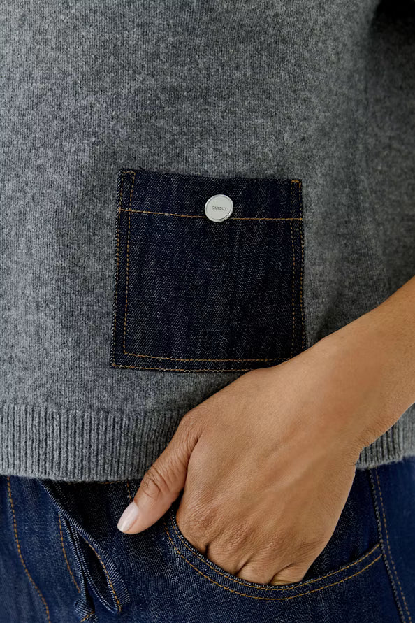 Close-up of a gray knit sweater with a dark blue denim patch pocket featuring a silver button, worn by a model with blue jeans.