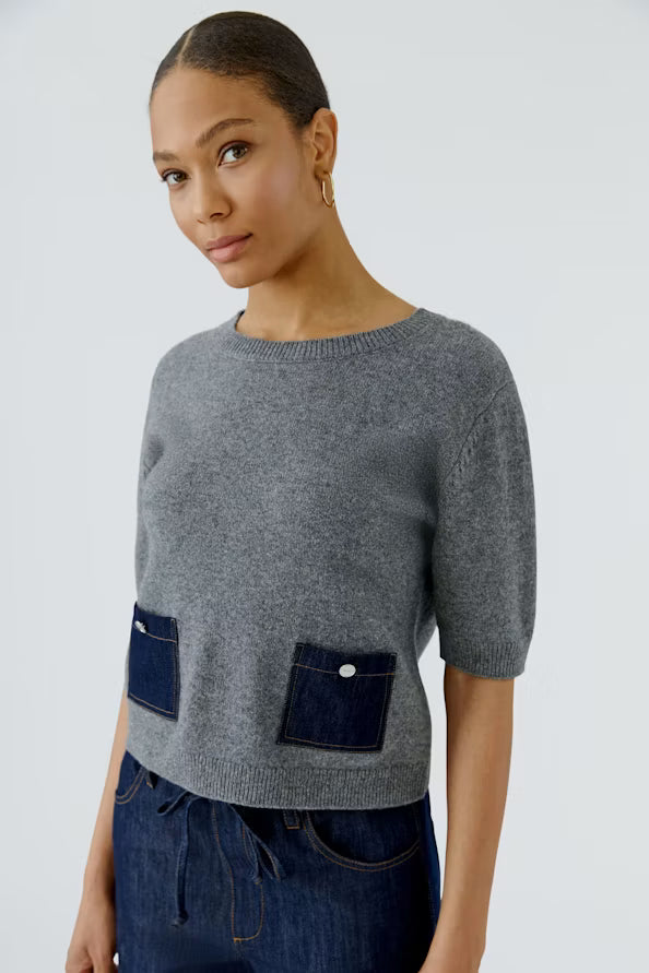A woman wearing a gray short-sleeve knit sweater with two dark blue patch pockets on the front, paired with dark blue jeans, against a light gray studio background.