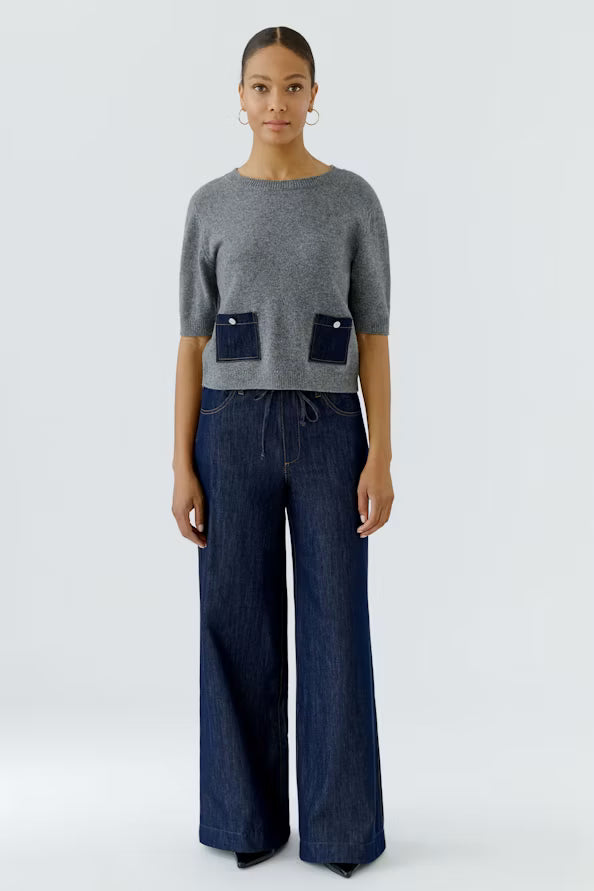 A model stands against a light gray studio background wearing a gray short-sleeve knit top with two dark blue patch pockets and dark blue wide-leg jeans.