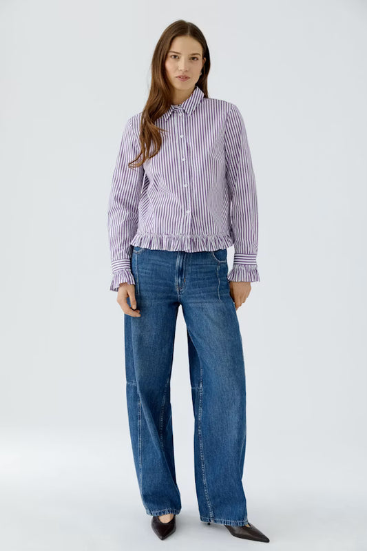 A woman modeling a lavender and white vertically striped button-down shirt with a ruffled hem, paired with wide-leg blue jeans and black pointed flats, against a light gray studio background.