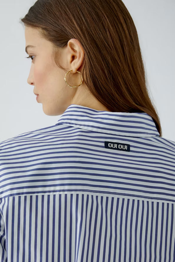 A close-up side profile of a woman wearing a blue and white vertically striped button-down shirt and a gold hoop earring. The brand name 'OUI OUI' is visible on a small black label sewn onto the back of the shirt.