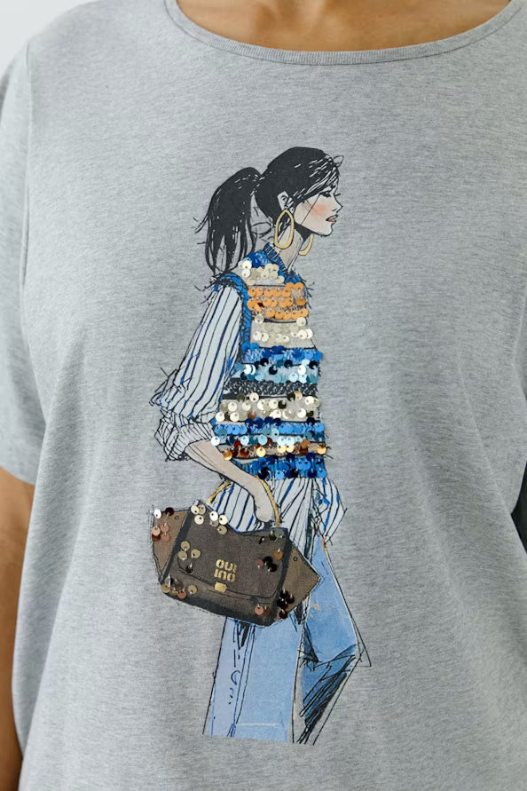 A close-up of a light gray heather t-shirt featuring a fashion illustration of a woman in a striped top, blue jeans, and a brown handbag, with the words 'FASHION BAG' printed on the graphic.
