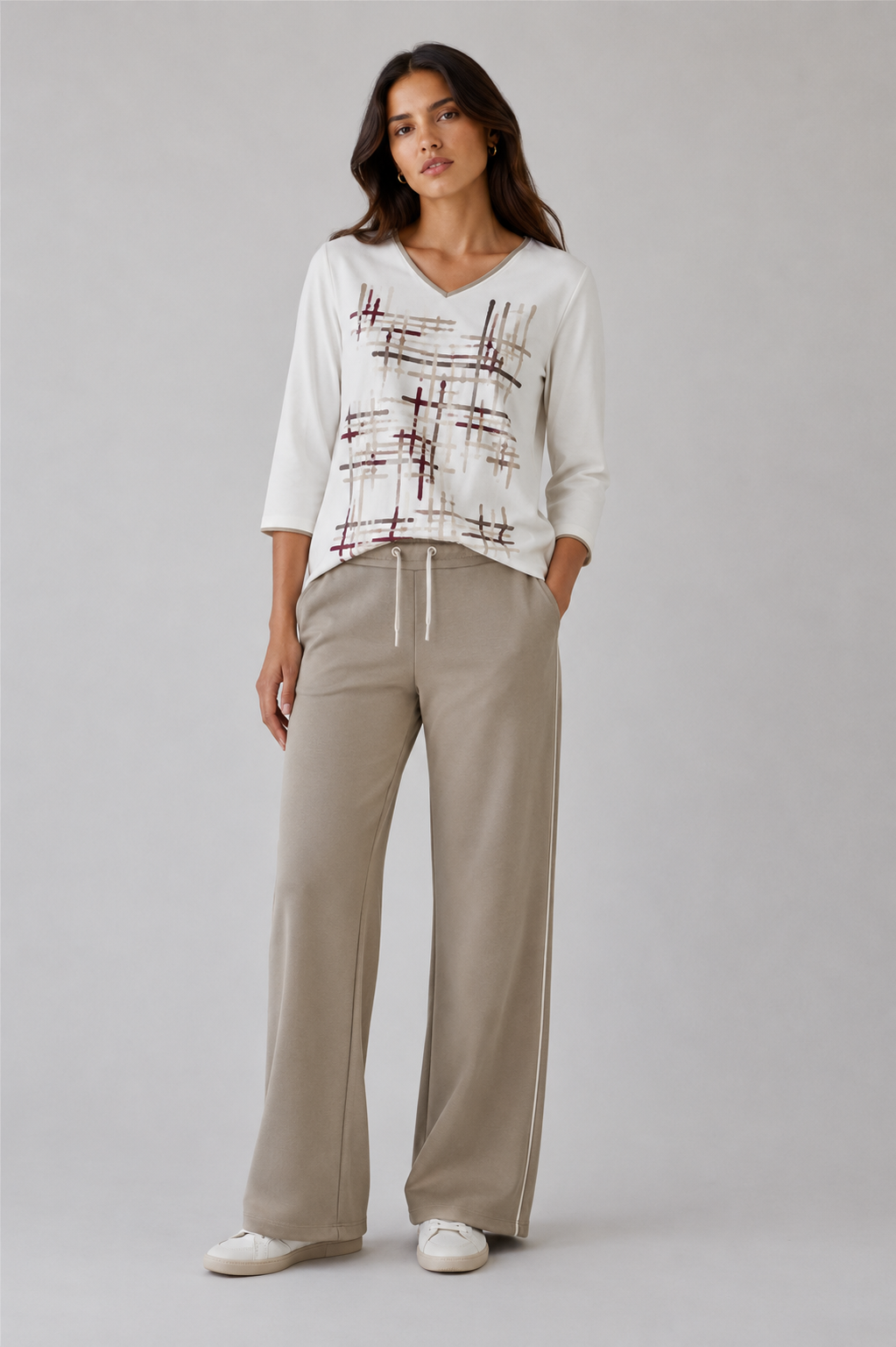 A woman modeling a white V-neck knit top with a brown abstract grid pattern on the front, paired with beige wide-leg drawstring trousers and white sneakers, against a light gray studio background.
