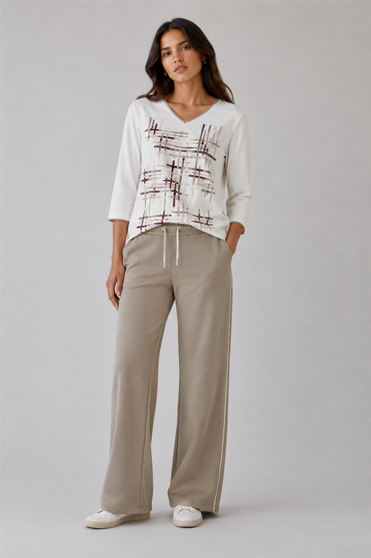 A woman modeling a white V-neck knit top with a brown abstract grid pattern on the front, paired with beige wide-leg drawstring trousers and white sneakers, against a light gray studio background.