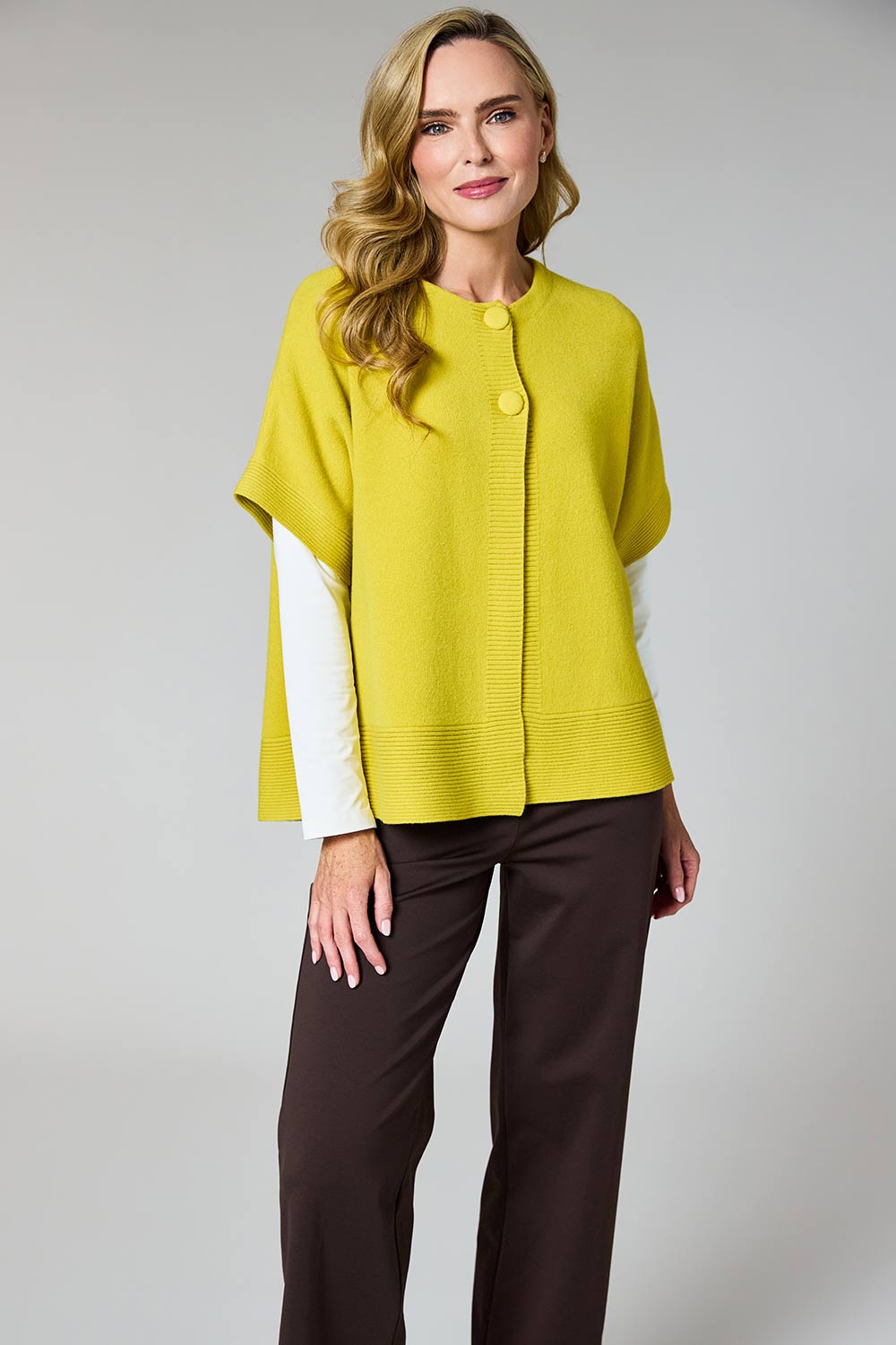 A woman modeling a bright yellow knit cape cardigan with white sleeve panels and dark brown trousers, shot against a neutral gray studio background.
