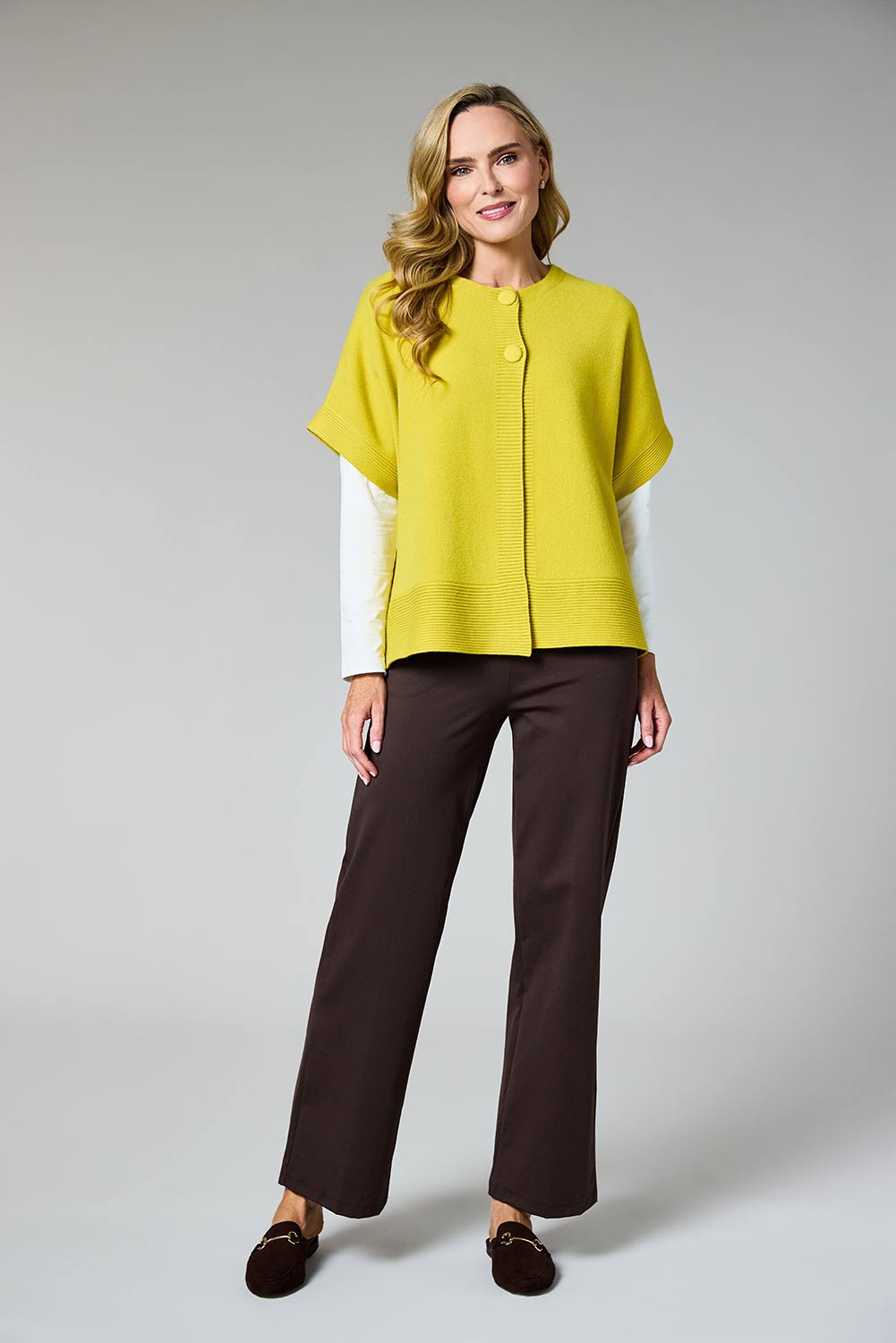 A woman modeling a bright yellow short-sleeve knit poncho-style top over a white long-sleeve shirt, paired with dark brown wide-leg trousers and dark brown loafers, against a light gray studio background.