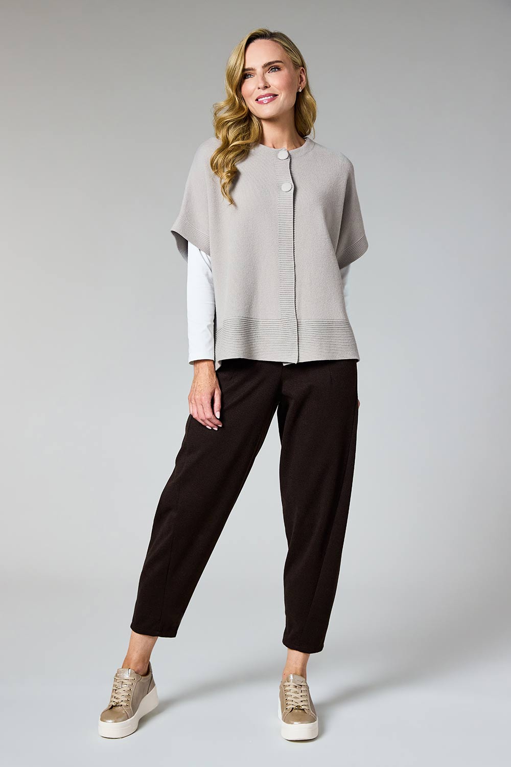 A woman modeling a light gray knit poncho-style top over a white long-sleeve shirt, paired with dark brown cropped pants and beige sneakers, against a light gray studio background.