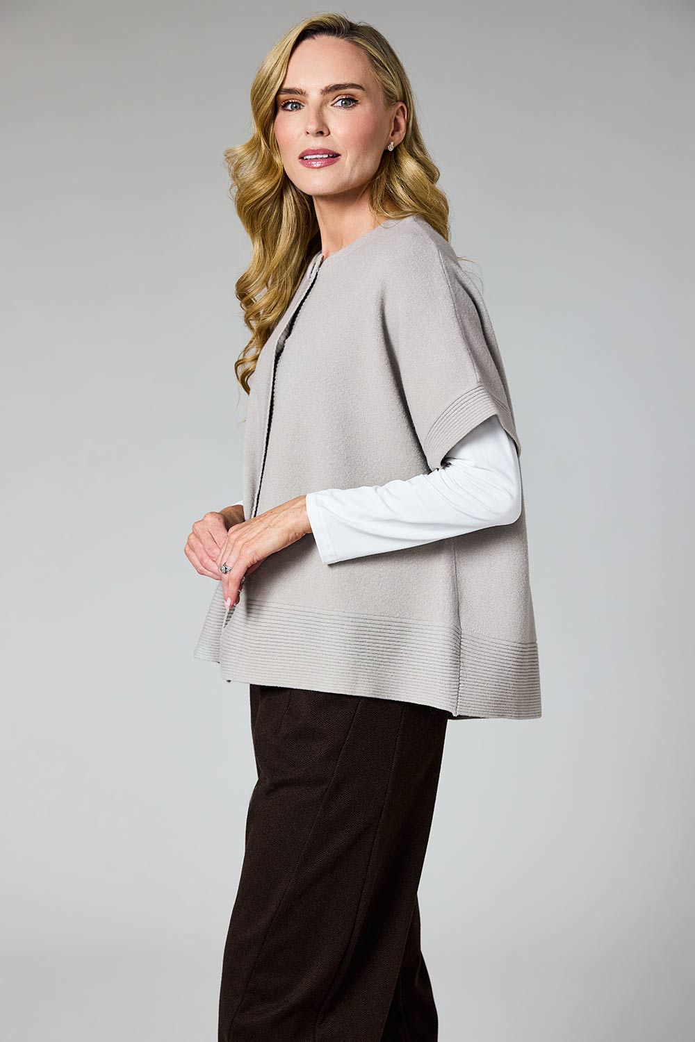 A woman modeling a light gray knit poncho-style cardigan over a white long-sleeve top and dark brown pants, photographed against a neutral light gray studio background.