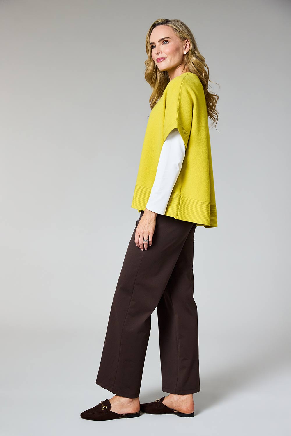 A woman modeling a bright yellow cape-sleeve top over a white long-sleeve shirt, paired with dark brown wide-leg trousers and dark brown loafers, against a light gray studio background.