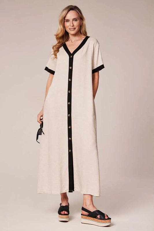 A woman modeling a cream-colored maxi dress with black trim along the V-neckline, sleeve cuffs, and front button placket. She is wearing black platform sandals against a solid beige studio background.