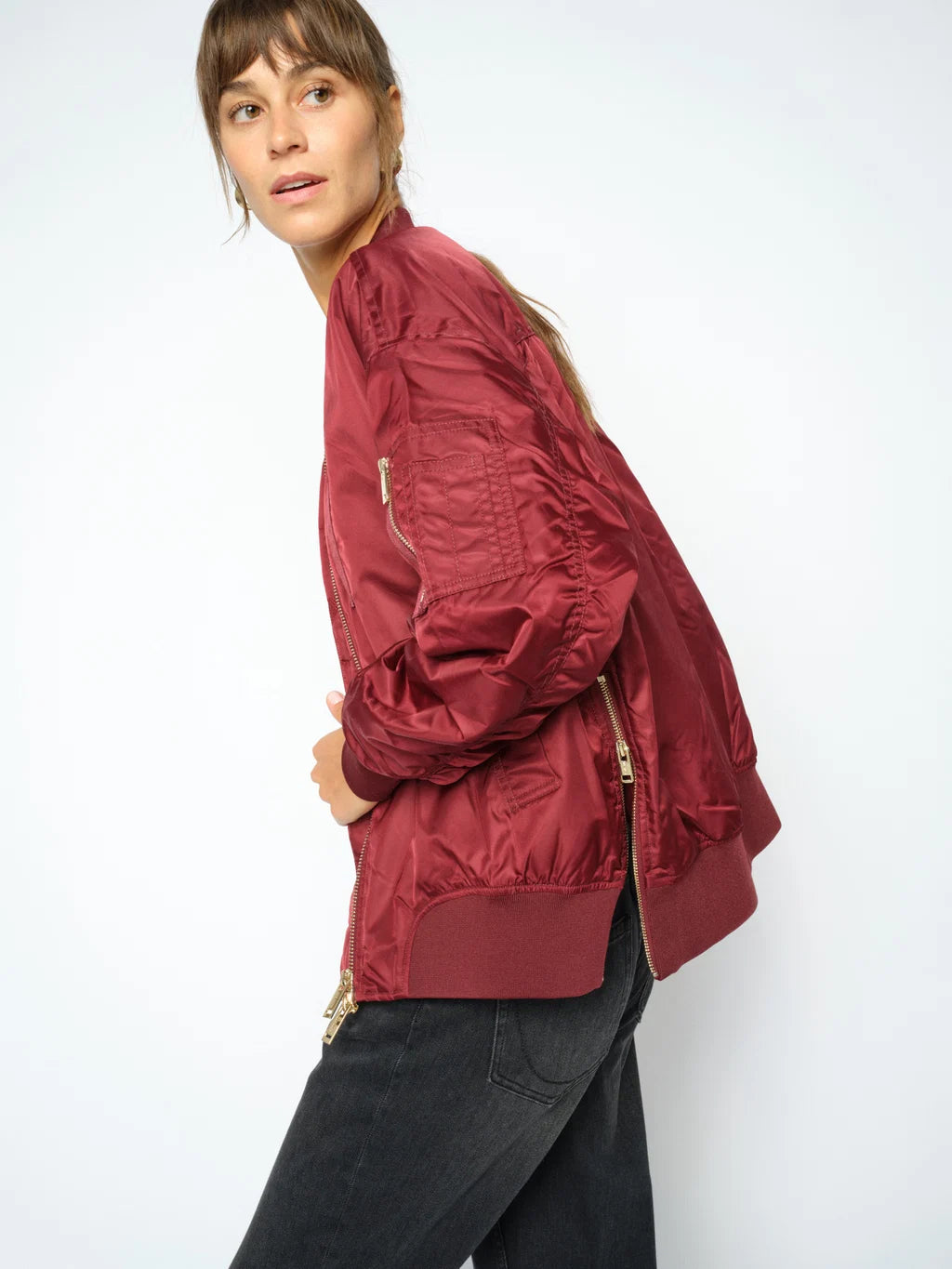 A woman wearing a burgundy satin bomber jacket with gold-tone zippers, paired with black jeans, photographed against a clean white studio background.