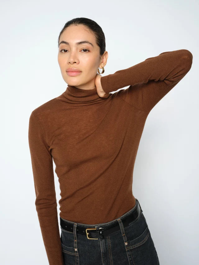 A woman modeling a fitted, long-sleeve brown turtleneck top paired with dark blue jeans and a black belt, set against a plain light gray studio background.