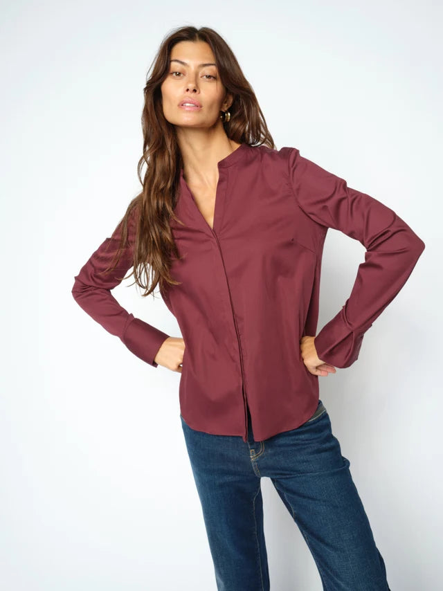 A woman modeling a long-sleeve button-up blouse in a deep burgundy or maroon shade, paired with dark blue jeans, against a plain light gray studio background.