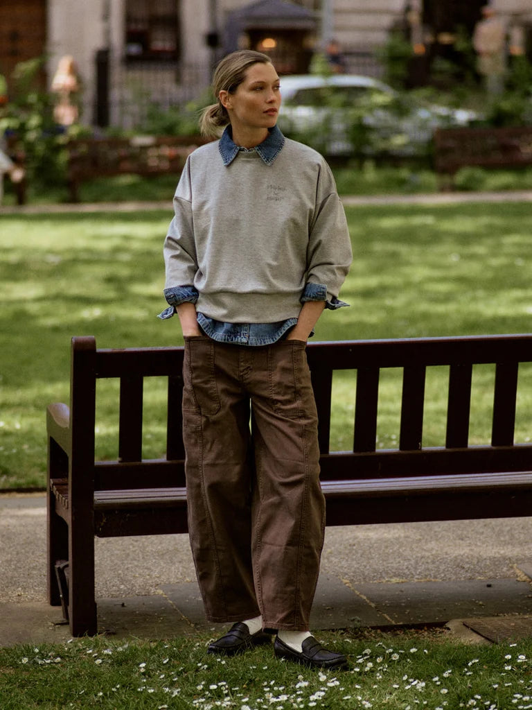 A woman stands outdoors on a bench in a park-like setting, wearing a light gray sweatshirt over a blue collared shirt, brown wide-leg pants, and dark loafers.