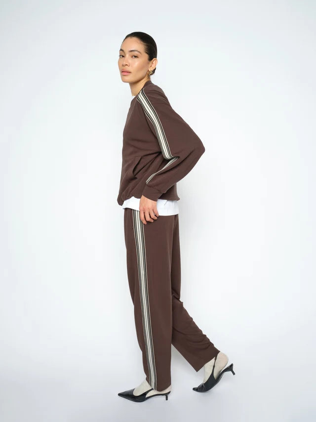 A model wearing a matching brown knit tracksuit with double cream stripes down the sleeves and legs, styled with a white t-shirt, white undershirt, and black-and-white pointed-toe heels, set against a plain light gray studio background.