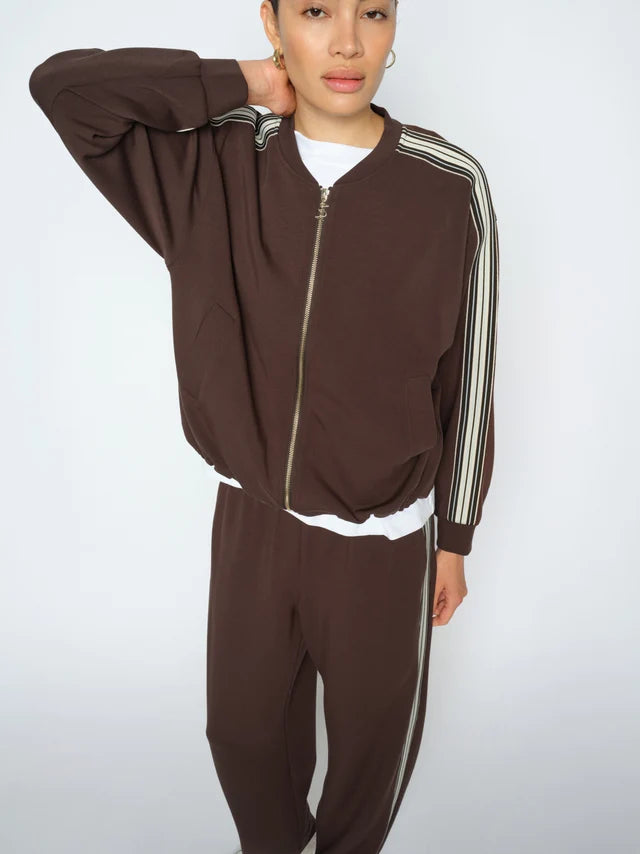 A model wearing a matching brown zip-up track jacket and jogger pants set with white and black stripes down the sleeves and legs, styled over a white t-shirt against a light gray background.