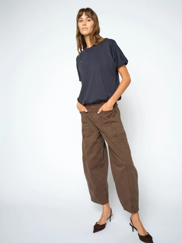 A woman modeling a loose navy blue short-sleeve top and brown wide-leg cropped pants with dark brown pointed-toe heels, standing against a light gray studio background.