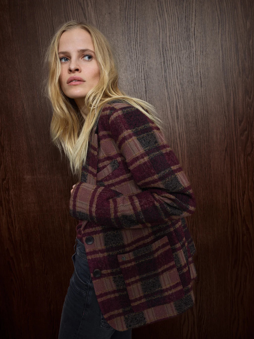 A blonde woman wearing a burgundy and brown plaid wool-blend blazer over a dark top, posed against a dark wood-paneled background.