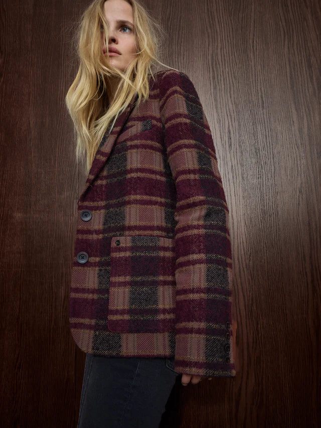 A woman wearing a long-sleeve plaid blazer in burgundy, brown, and beige tones, paired with dark jeans, posed against a dark wood-paneled background.