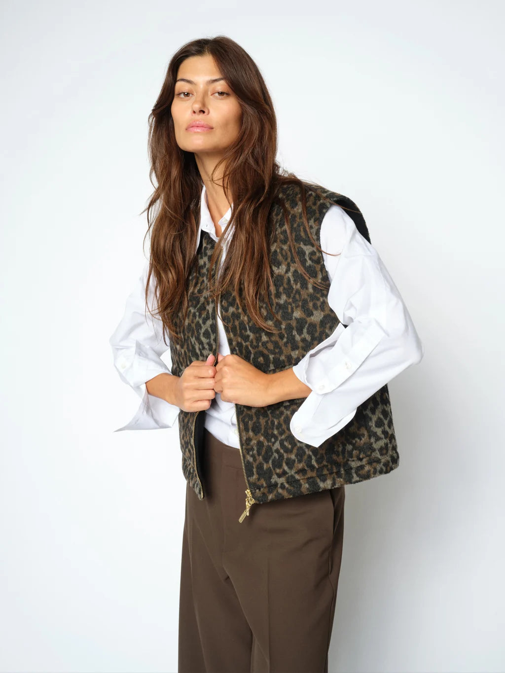 A woman modeling a leopard-print vest over a white collared shirt and brown trousers, standing against a plain light gray studio background.
