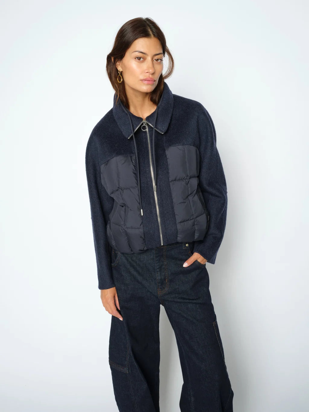 A woman modeling a dark navy blue jacket with a quilted puffer chest panel and a silver zipper, paired with dark jeans, against a plain light gray studio background.