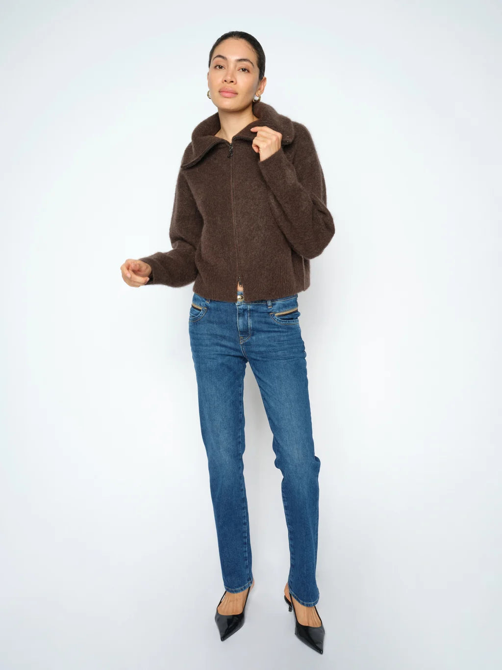 A woman modeling a brown fuzzy cropped jacket with a high collar, paired with blue jeans and black pointed-toe heels, against a plain light gray studio background.