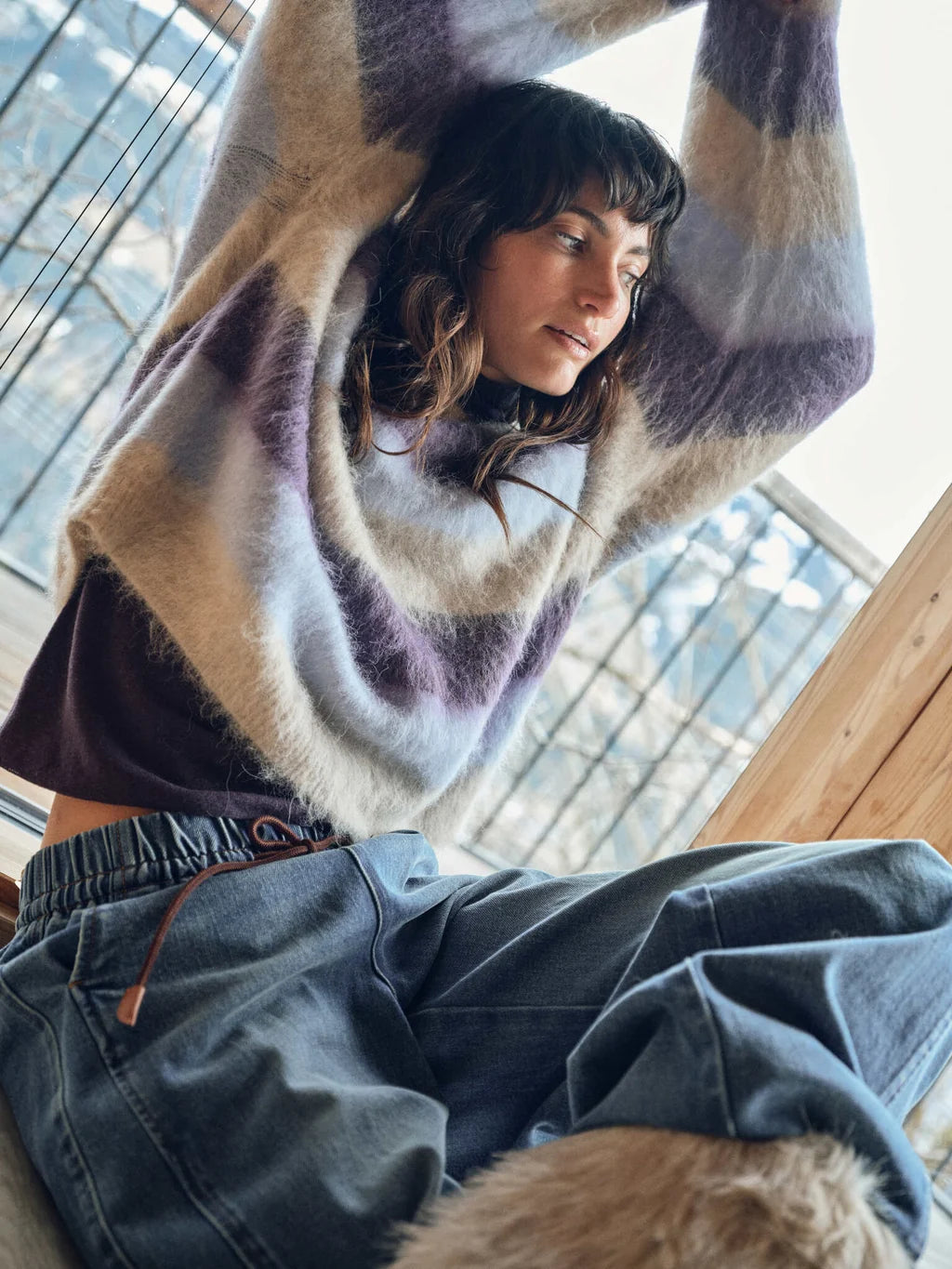 Person wearing a patterned sweater and jeans sitting indoors with a view of a cityscape through large windows.