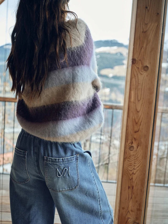 A person viewed from behind wearing a fuzzy striped sweater with horizontal bands of cream, lavender, and gray, paired with light blue jeans, standing by a large window with a mountain view.